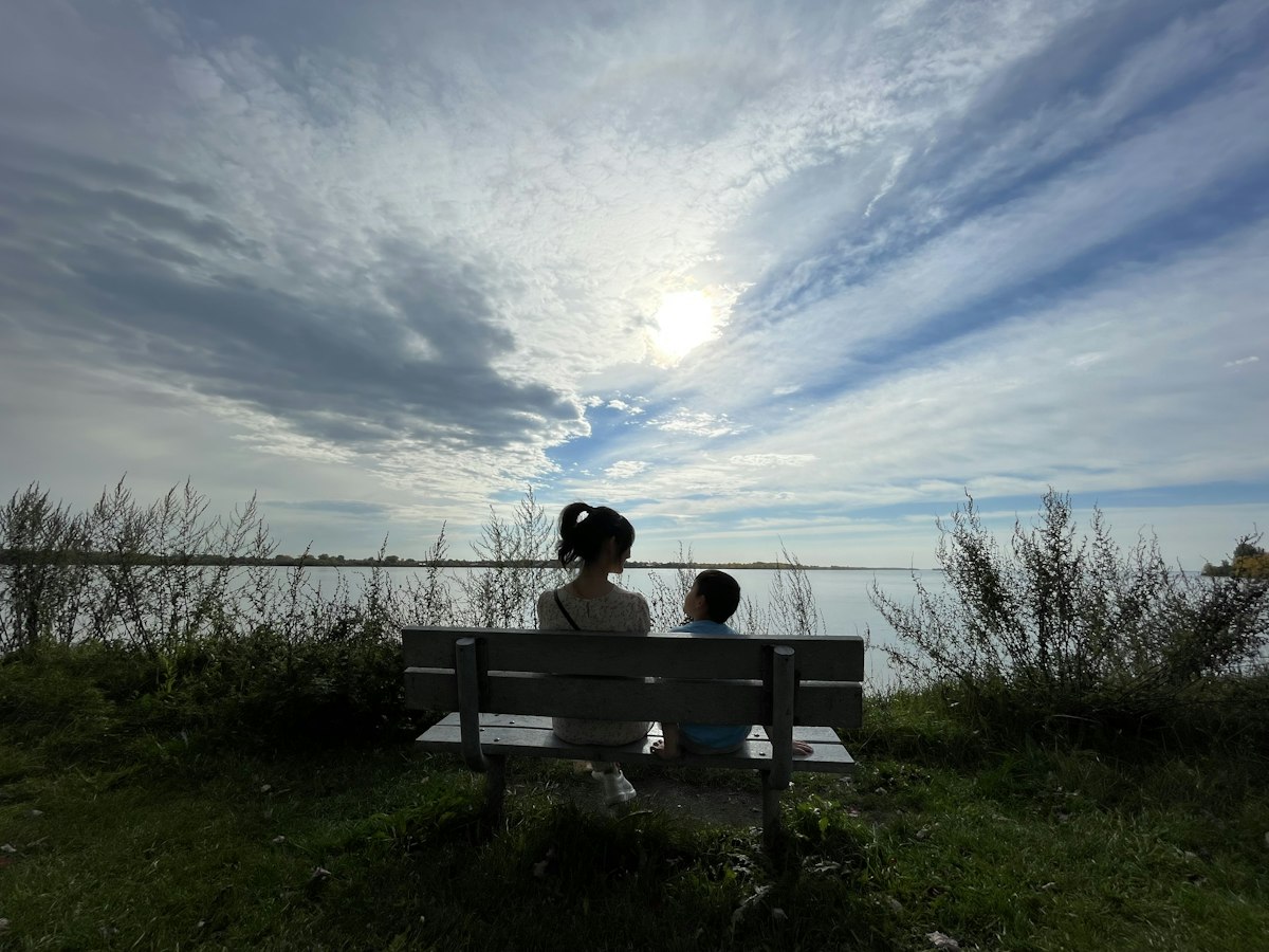 Eine Frau und ein Kind sitzen im Gespräch auf einer Bank mit Blick auf einen See, von hinten und im Gegenlicht, Gesichter nicht erkennbar
